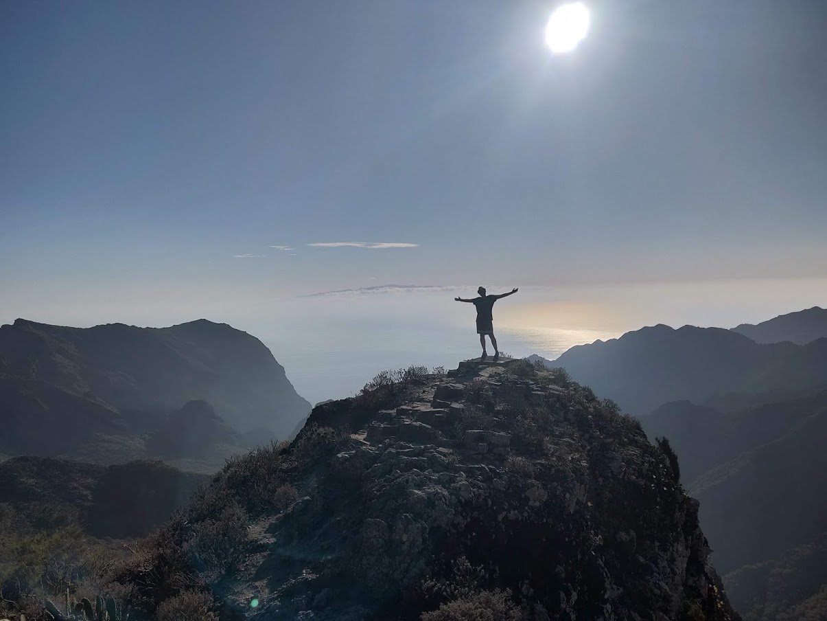 Silhouette di una persona a braccia aperte sulla cima di una montagna, in controluce sul mare